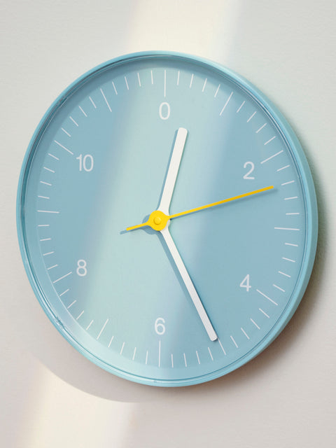 HAY Wall Clock, Blue, recycled ABS wall clock, light blue face with white hands, mounted on a pale wall in raking daylight
