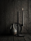 Black Museum Candle Holder with lit candle against dark textured wood backdrop, paired with ceramic vase.