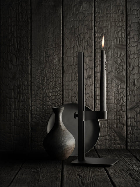 Black Museum Candle Holder with lit candle against dark textured wood backdrop, paired with ceramic vase.