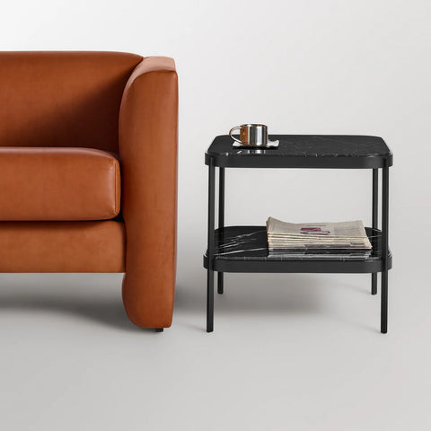 Blu Dot ComeHither Side Table in black marble styled beside a cognac leather sofa — two-tier design with marble top shelf holding a brass cup and lower shelf stacked with magazines