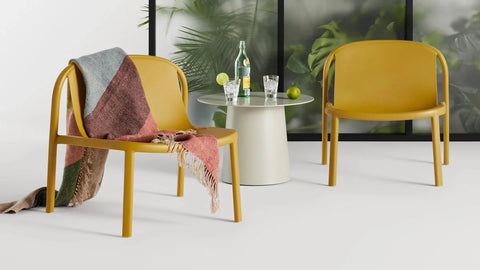 Blu Dot Decade Lounge Chairs in mustard recycled plastic, styled indoors with a round side table, sparkling water, limes, and a patterned throw draped over one chair