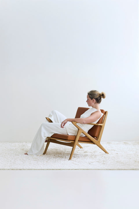 A woman seated in a DK3 Z Chair in oiled oak with cognac leather on a white rug