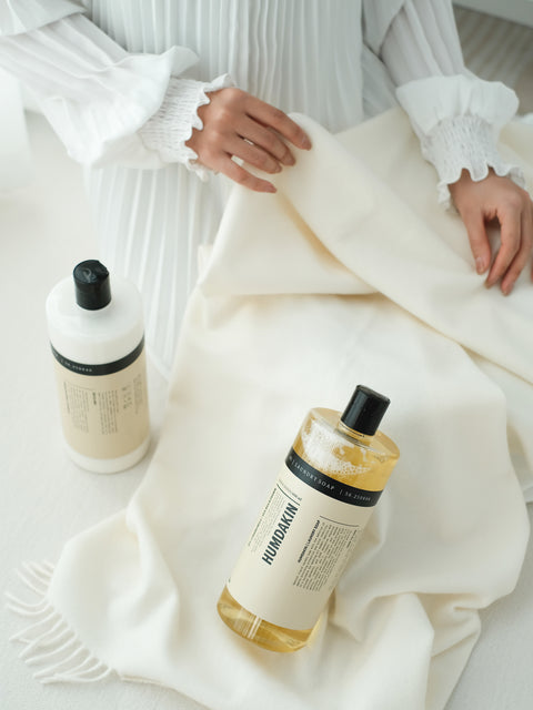 Humdakin 01 Fabric Softener, Chamomile & Sea Buckthorn, liquid fabric softener, softener and laundry soap bottles on a white knitted throw being folded by hand