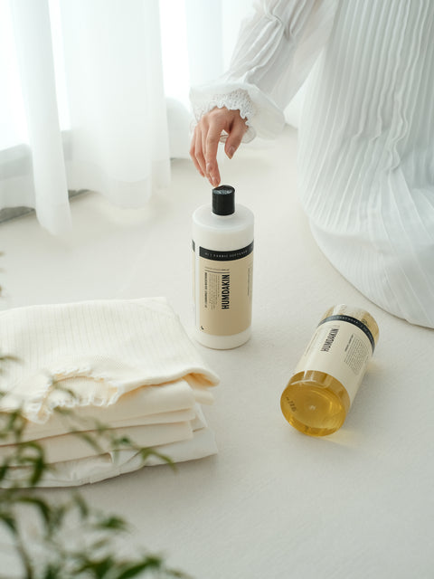 Humdakin 01 Laundry Soap, Chamomile & Sea Buckthorn, liquid laundry detergent, a hand reaching for the bottle beside folded white laundry and a sprig of greenery