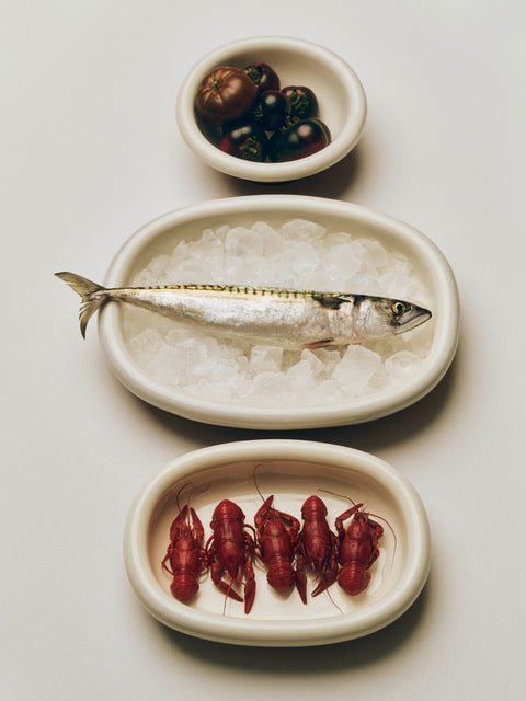 HAY Barro Oval Dish, Large, Off-White, glazed terracotta oval serving dish, holding a whole mackerel on ice beside dishes of crayfish and tomatoes