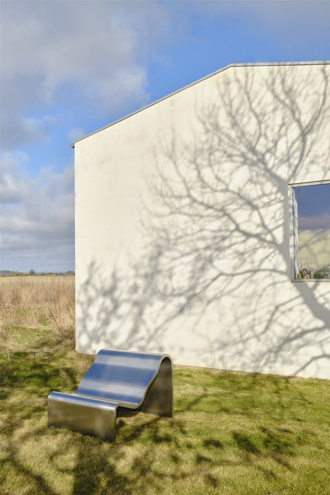 Muuto Dream View Bench in brushed steel outdoors beside a modern white house with tree shadows