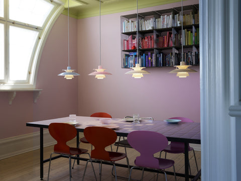 Four Louis Poulsen PH 5 Opal pendant lights in a row above a dining table with red and violet chairs, in a pink room with an arched window