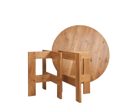 Frama Farmhouse Trestle Table in Natural Oak, round top standing on edge behind two open trestles with the parts not yet joined