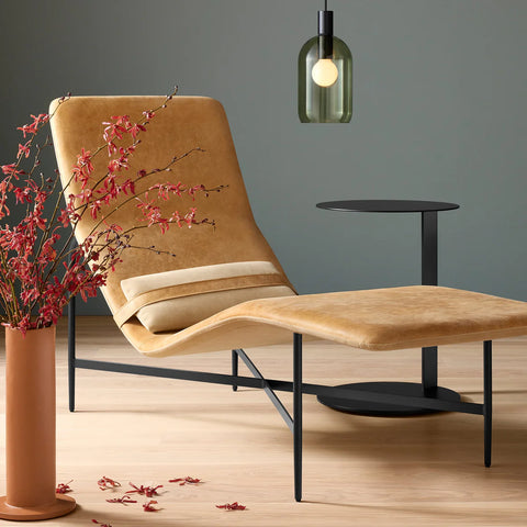 Blu Dot Deep Thoughts Leather Chaise in camel aniline leather with coordinating camel velvet lumbar pillow on black powder-coated steel base, styled beside a terracotta vase with red branches and a green glass pendant light
